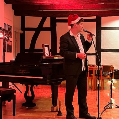 Daniel Wahren mit Nikolausmütze auf dem Kopf steht vor dem Flügel mit Mikrophone in der Hand und singt.